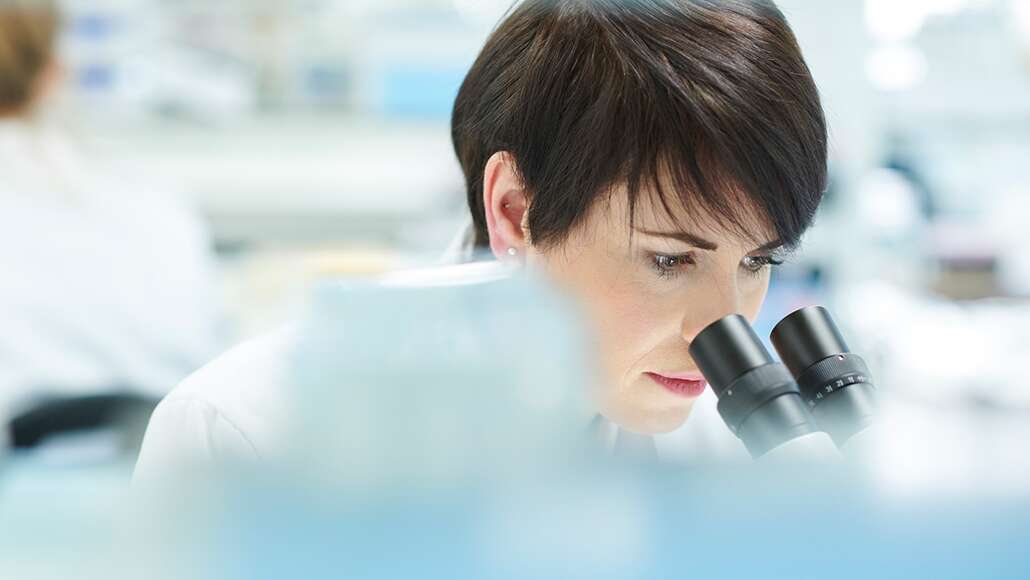 Female scientist in her lab looking into a microscope with another scientist in the background.